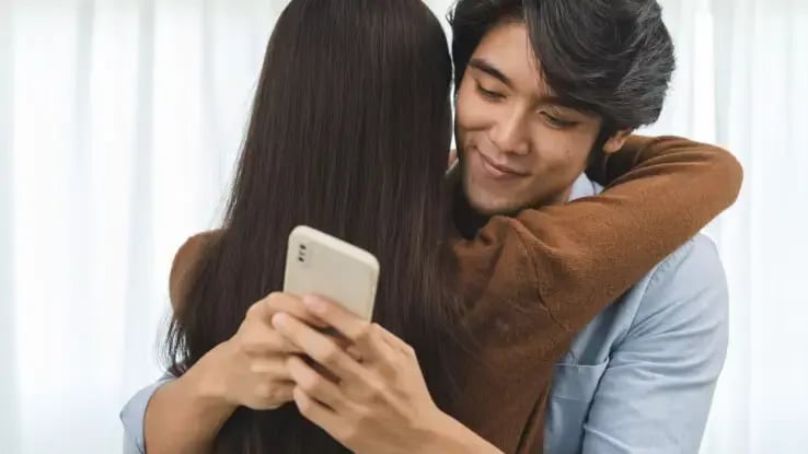 A man checking his phone while hugging his partner.